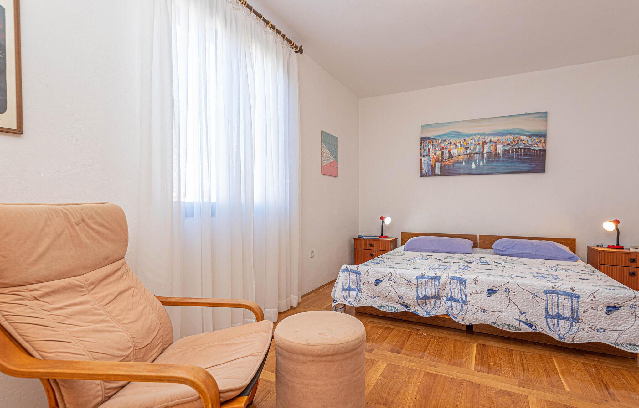 A bedroom with a bed, a chair, a round ottoman, a painting, and a nightstand with a lamp.