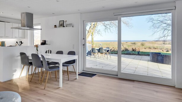 A modern kitchen with white cabinets, a dining area with a white table and grey chairs, and a large sliding glass door leading to an outdoor patio with a view of a field and trees. - Cozy home in Hadsund with WiFi (Hadsund)
