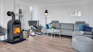 A modern living room with a wood-burning stove, a grey sofa, a rocking chair, and a small table with a vase of flowers. - Cozy home in Hadsund with WiFi (Hadsund)
