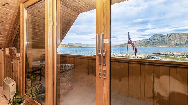 A wooden cabin with a glass door leading to a balcony overlooking a lake and mountains.