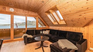 A cozy cabin interior with a wooden ceiling, large windows offering a lake view, a dark-colored sofa, and a small round table.