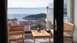 Un balcon agrémenté de meubles en bois, une table avec deux verres de jus d’orange et une vue sur une ville côtière et la mer.