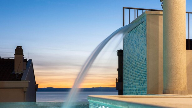 Une piscine agrémentée d’un élément décoratif aquatique, un mur carrelé et une vue sur le plan d’eau au coucher du soleil.