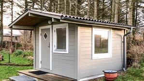 Ein kleines, eingeschossiges Gartenhaus mit Tür und zwei Fenstern, eine Holzrampe führt zur Tür, und ein roter Eimer liegt in der Nähe des Eingangs.