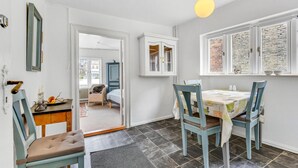 Et værelse med et spisebord, stole, et skab, et spejl og et vindue med udsigt til en murstensbygning.