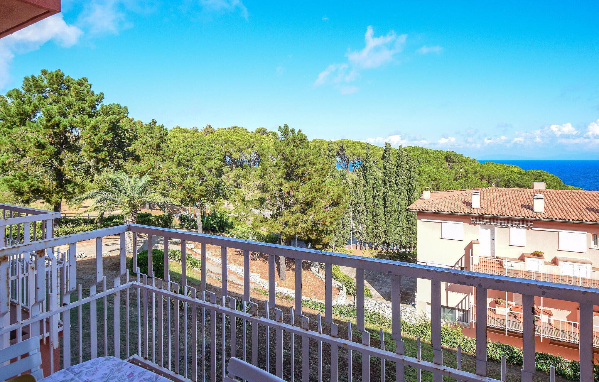 A balcony with a white railing overlooking a coastal area with trees and a building with a red roof.