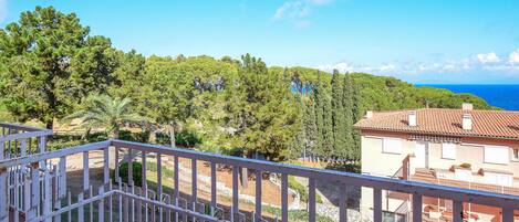 A balcony with a white railing overlooking a coastal area with trees and a building with a red roof.