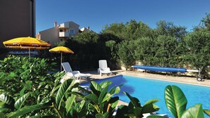 Un espace piscine avec des chaises longues et des parasols, entouré de verdure et avec un bâtiment en arrière-plan.