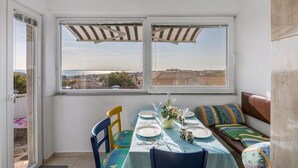 A dining area with a table set for two, a bench with cushions, and a view of the city through large windows. - Nice apartment in Barbariga with WiFi (Barbariga)