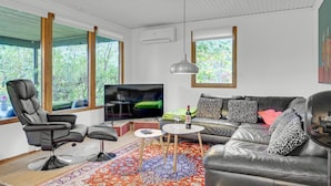 Un salon moderne avec un canapé en cuir, un fauteuil en cuir noir, un téléviseur et une grande fenêtre donnant sur la forêt.