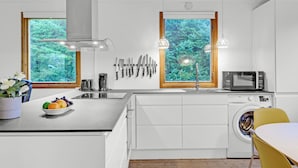 Une cuisine moderne dotée d’un îlot central, d’un lave-linge encastré et offrant une vue sur la verdure grâce à de grandes fenêtres.