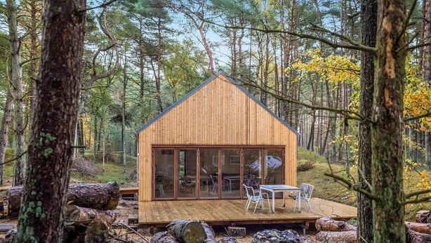 A wooden cabin with a large glass door, a deck with chairs, and logs in the foreground. - Nice home in Nexø with WiFi (Nexø)