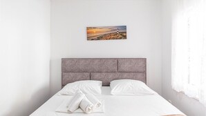 Un lit bien fait, avec des draps blancs et deux oreillers, une tête de lit grise et un tableau représentant un coucher de soleil sur une plage avec un phare.