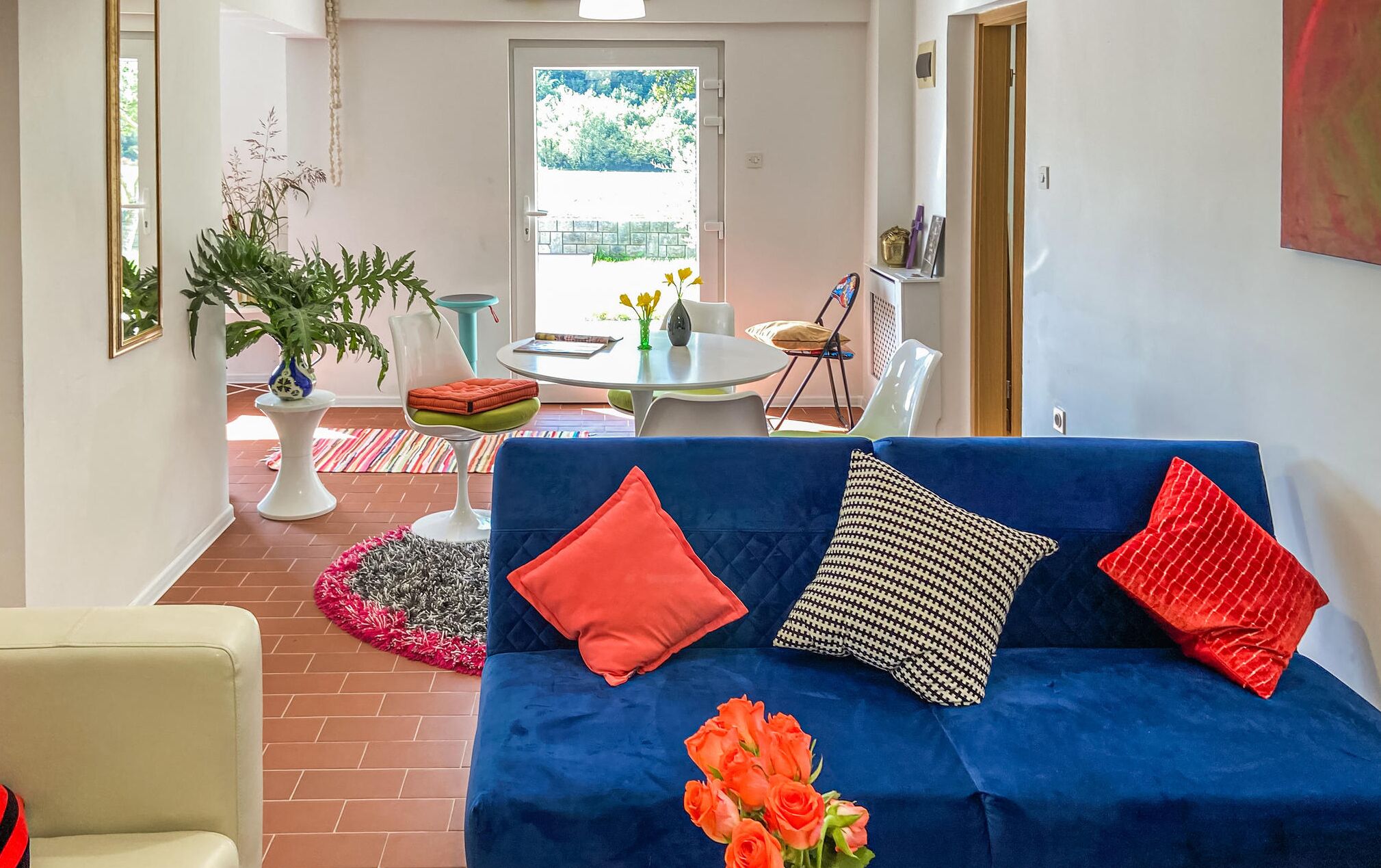 Un salon avec un canapé bleu, des coussins à motifs rouges et noirs, une plante en pot et un coin repas comprenant une table et des chaises.