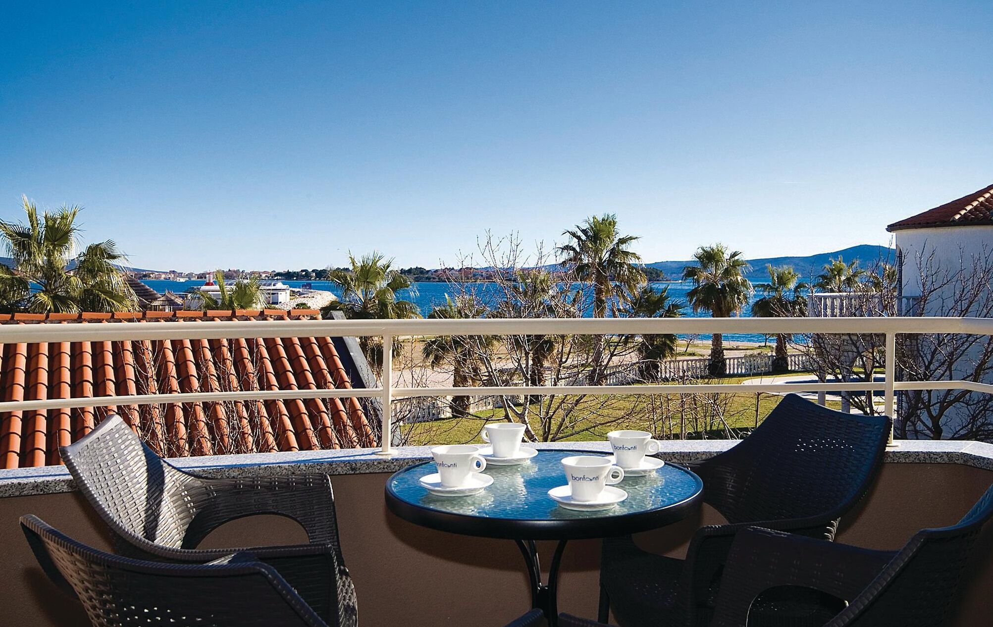 A terrace with a table set for four, overlooking a coastal area with palm trees and a view of the sea.