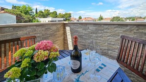 Una mesa con una botella de Prosecco, copas y un ramo de flores en una terraza con paredes de piedra y vistas a edificios.