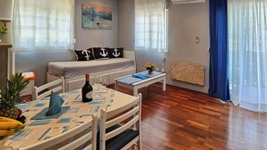 Una habitación acogedora con suelo de madera, una mesa de comedor con sillas, un sofá con almohadones de temática náutica, un cuadro en la pared y una ventana con cortinas.