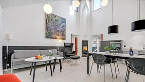 Ett modernt vardagsrum med en grå soffa, ett runt soffbord och en matplats med ett vitt bord och svarta stolar. En öppen spis syns i bakgrunden, och en stor tavla hänger på väggen.