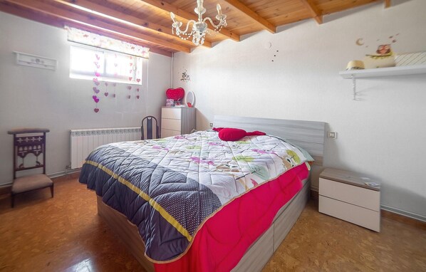 Un dormitorio con suelo de madera, una cama con colcha, una mesita de noche, una silla, un aparador, un radiador, un ventanal con adornos de corazón y una araña.