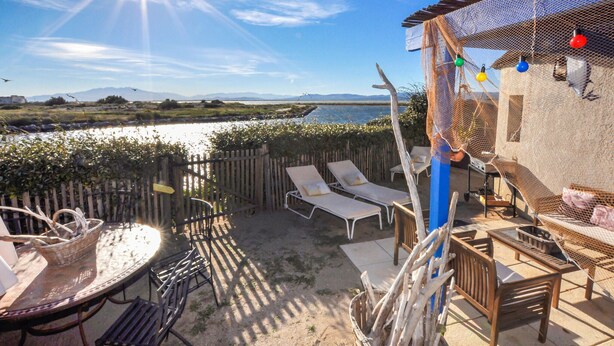 Eine Terrasse mit Blick auf ein Gewässer, Holzmöbel und eine Pergola mit Lichterketten.
