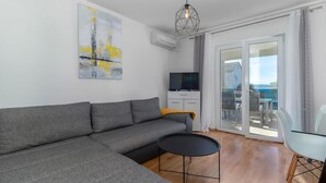 Ein modernes Wohnzimmer mit einem grauen Ecksofa, einem kleinen schwarzen Couchtisch, einem Flachbildfernseher auf einem weißen Schrank und einem Balkon mit Gartenmöbeln.