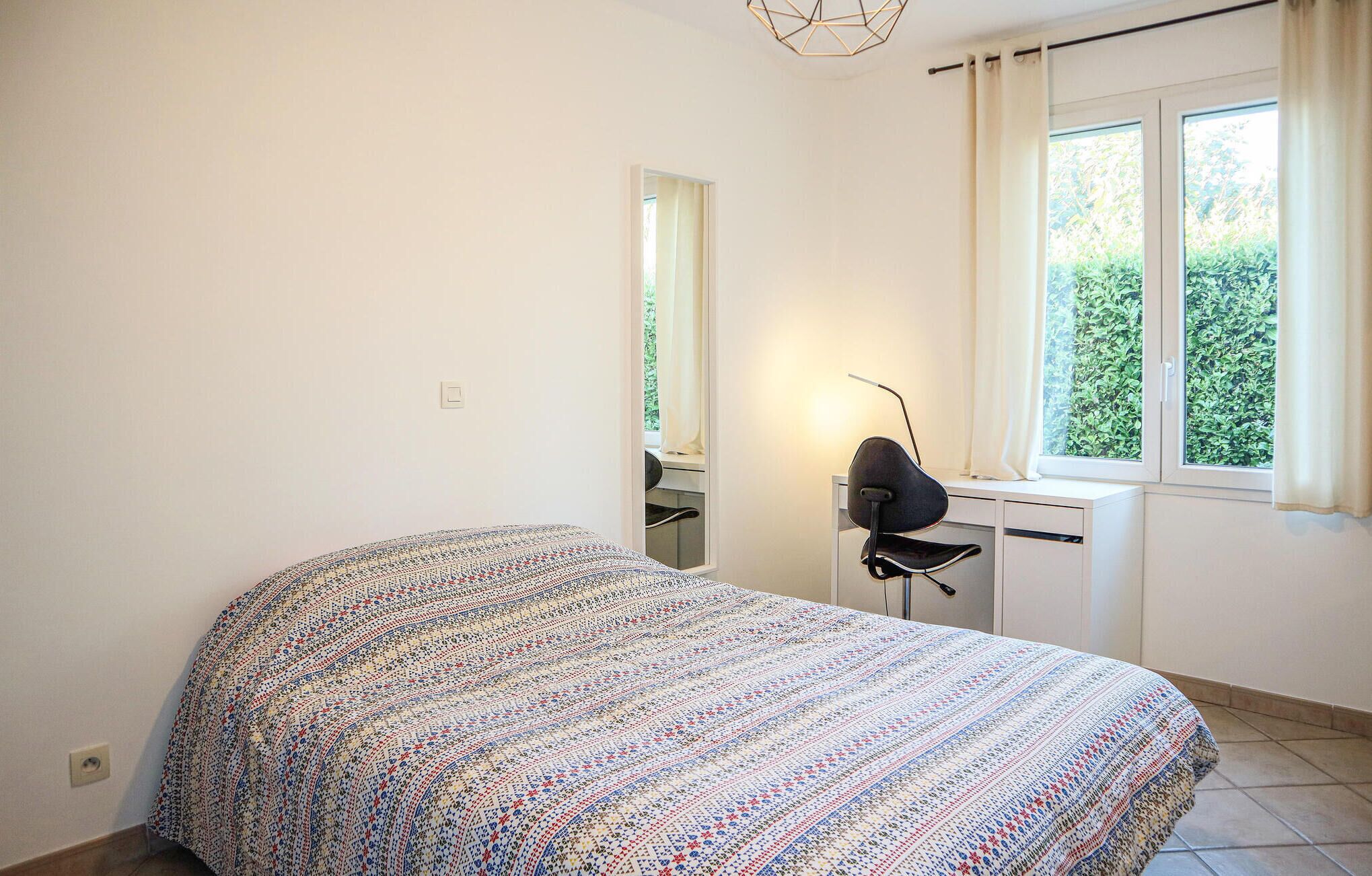 A bedroom with a patterned bedspread, a desk with a chair, a mirror, and a window with curtains.