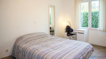 A bedroom with a patterned bedspread, a desk with a chair, a mirror, and a window with curtains.