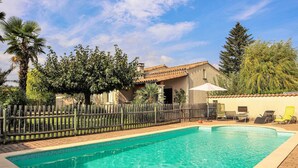 Una piscina con bordo piastrellato, circondata da una recinzione in legno, e un edificio con tetto in tegole sullo sfondo.