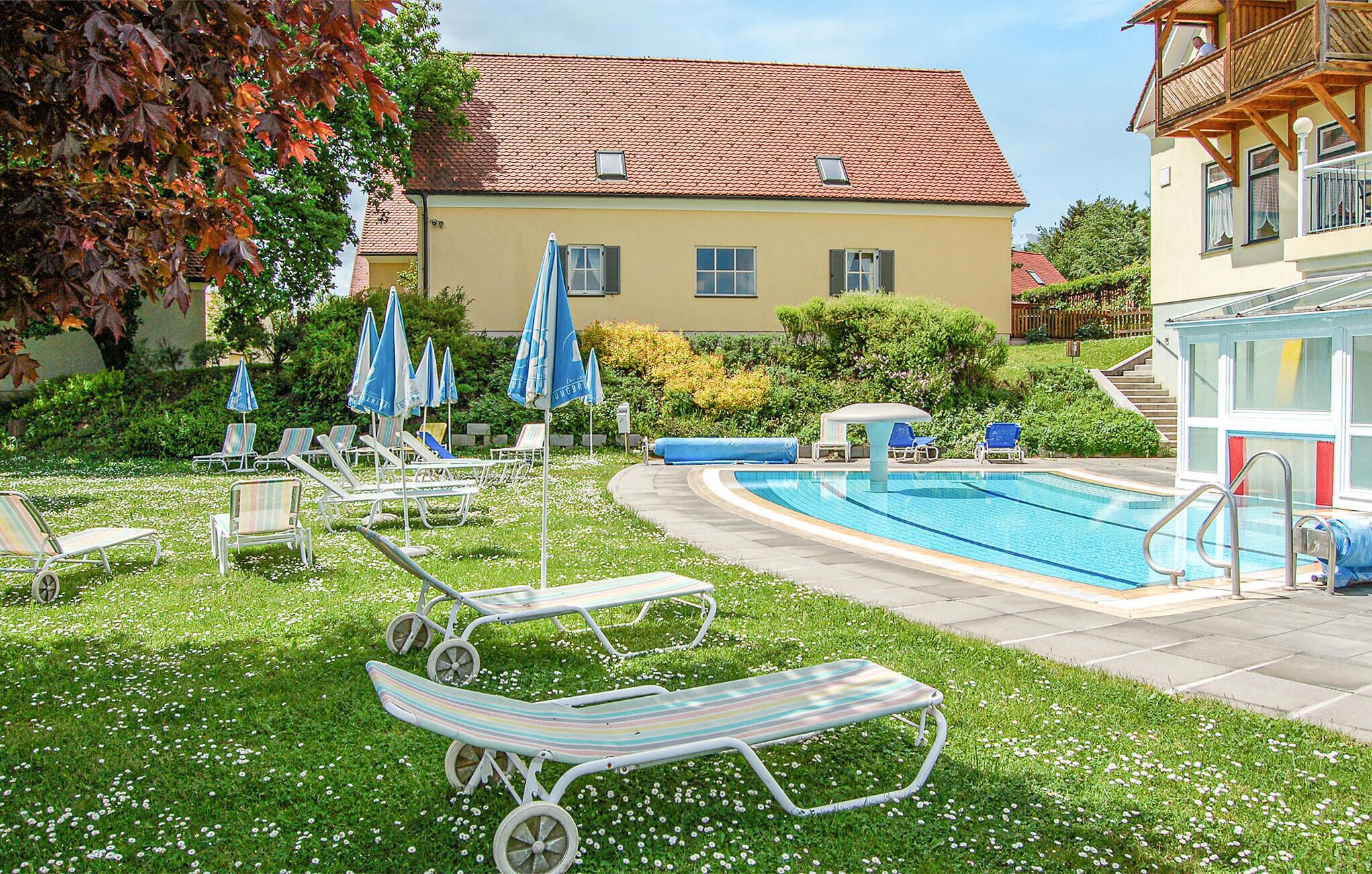 Ein Swimmingpool mit Liegestühlen und Sonnenschirmen, umgeben von einem gepflegten Rasen und einem Gebäude mit rotem Ziegeldach.