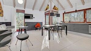 Une cuisine moderne avec un coin repas, un téléviseur fixé au mur et une grande fenêtre donnant sur une forêt.