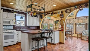 A kitchen with white cabinets, a central island with two chairs, a hanging pot rack, and a view of the outside through a window. - FABULOUS BEACH HOUSE- 7 Bedrooms,Heated Pool,12 BeachBadges,9 Car Driveway (Point Pleasant Beach)