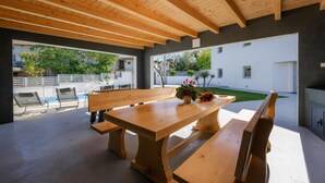 An outdoor patio with a wooden table and benches, a pool, and a view of a white building and greenery. - Beautiful home in Zuzici (Zuzici)