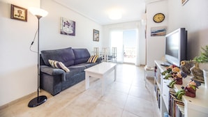 A modern living room with a grey sofa, a white coffee table, a floor lamp, and a TV on a stand. There are wall-mounted paintings and a clock. - 2 bedroom beach front apartment in Cabanes (Cabanes)