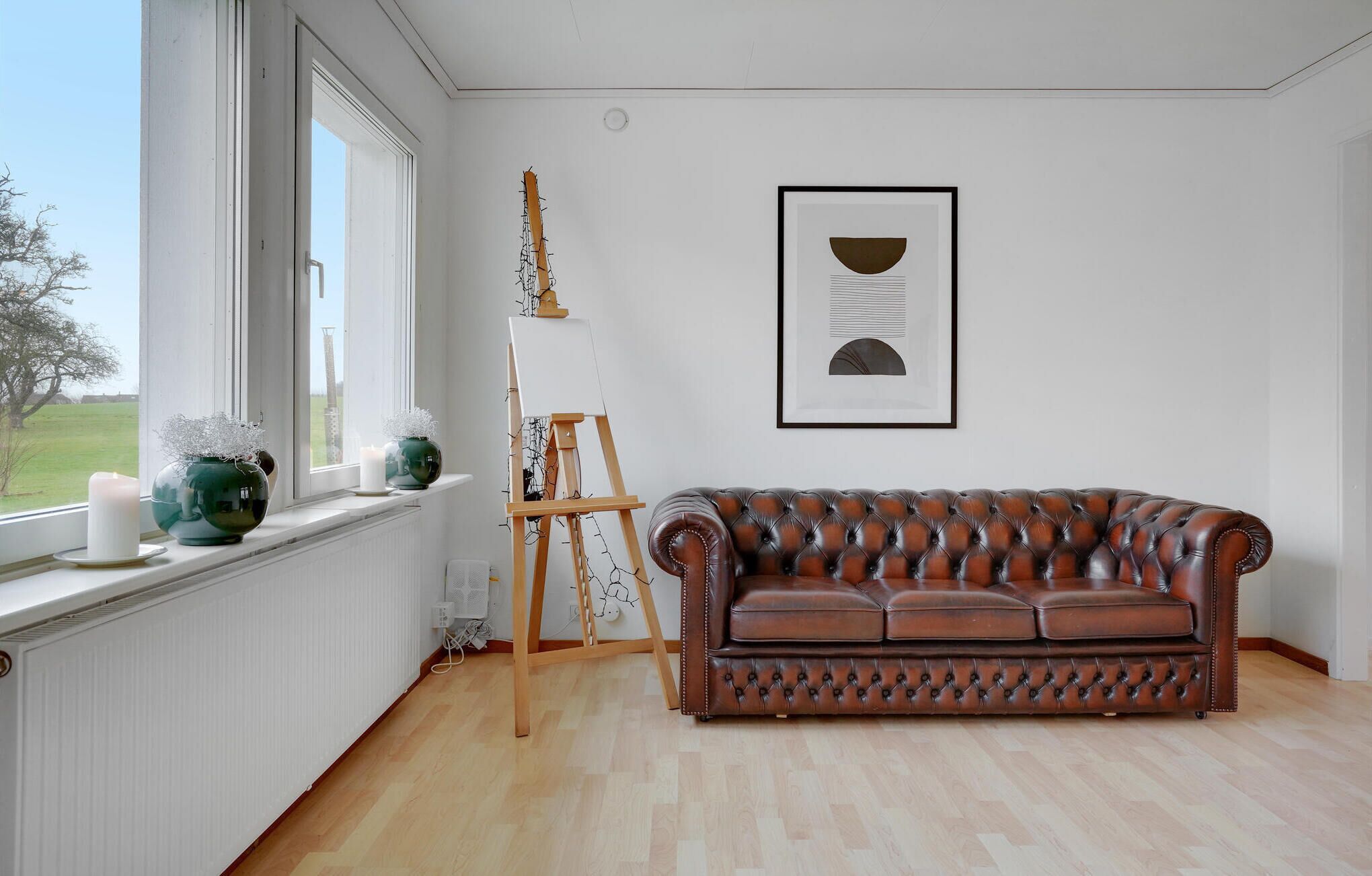 Une pièce avec un canapé en cuir marron, un tableau abstrait encadré et une fenêtre donnant sur un arbre et un immeuble.