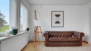 Une pièce avec un canapé en cuir marron, un tableau abstrait encadré et une fenêtre donnant sur un arbre et un immeuble.