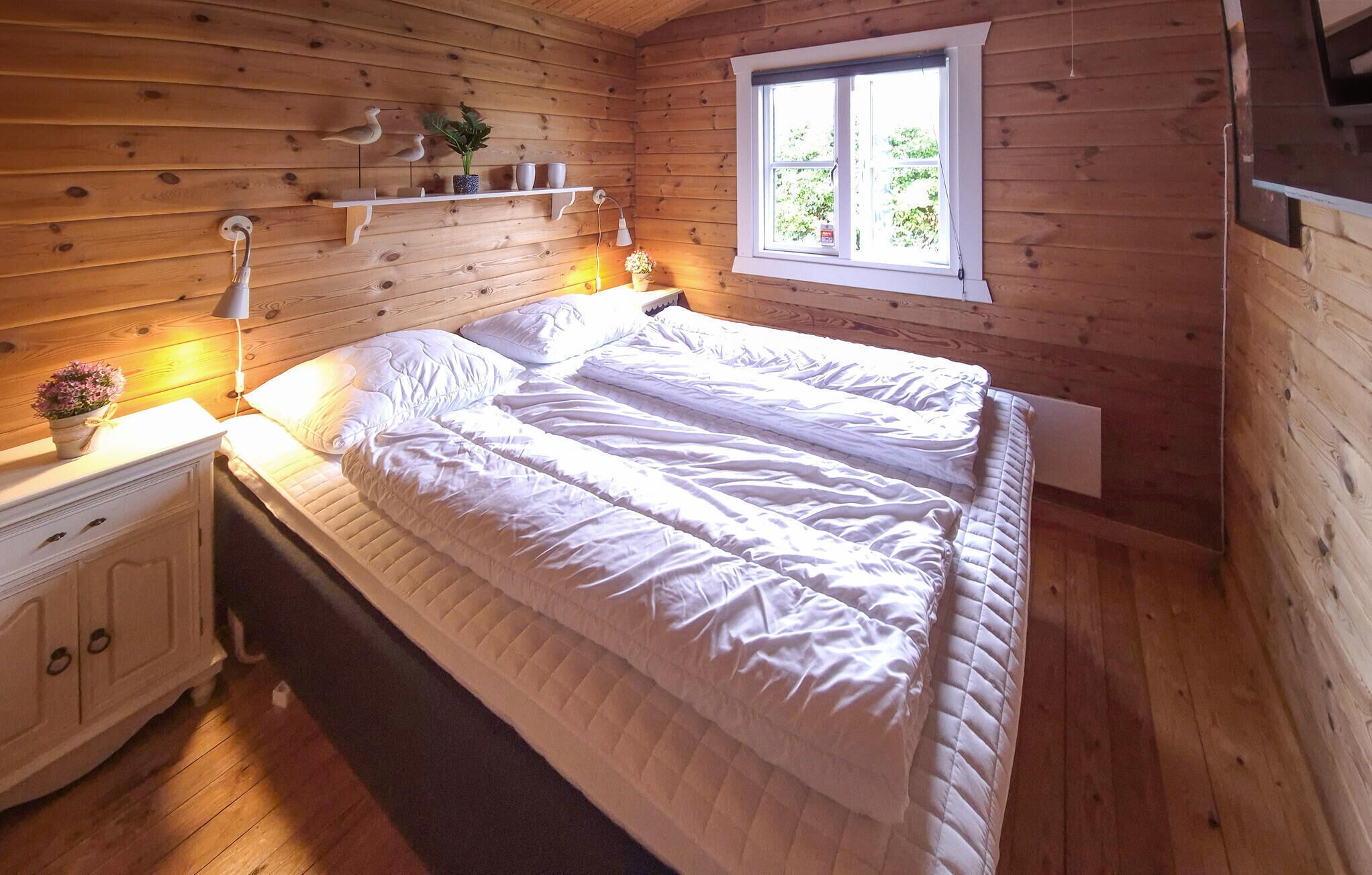 A wooden cabin bedroom with a bed, bedside table, lamp, and a window with a view of greenery.
