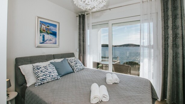 Een slaapkamer met een bed, kussens, een schilderij en uitzicht op zee door een groot raam.