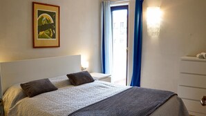 Um quarto com cama, mesa de cabeceira, cômoda, um quadro e uma janela com cortinas.