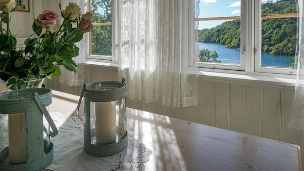 A table with a vase of roses and two candle holders, a lace doily, and a view of a lake through a window with lace curtains. - 4 bedroom beautiful home in Farsund (Farsund)