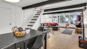 Un salon moderne avec un escalier, un coin repas avec une table dressée pour deux personnes, et une vue sur l’extérieur grâce à de grandes fenêtres.