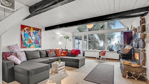 Un salon moderne avec un canapé d’angle gris, une cheminée en pierre et de grandes fenêtres donnant sur l’extérieur.