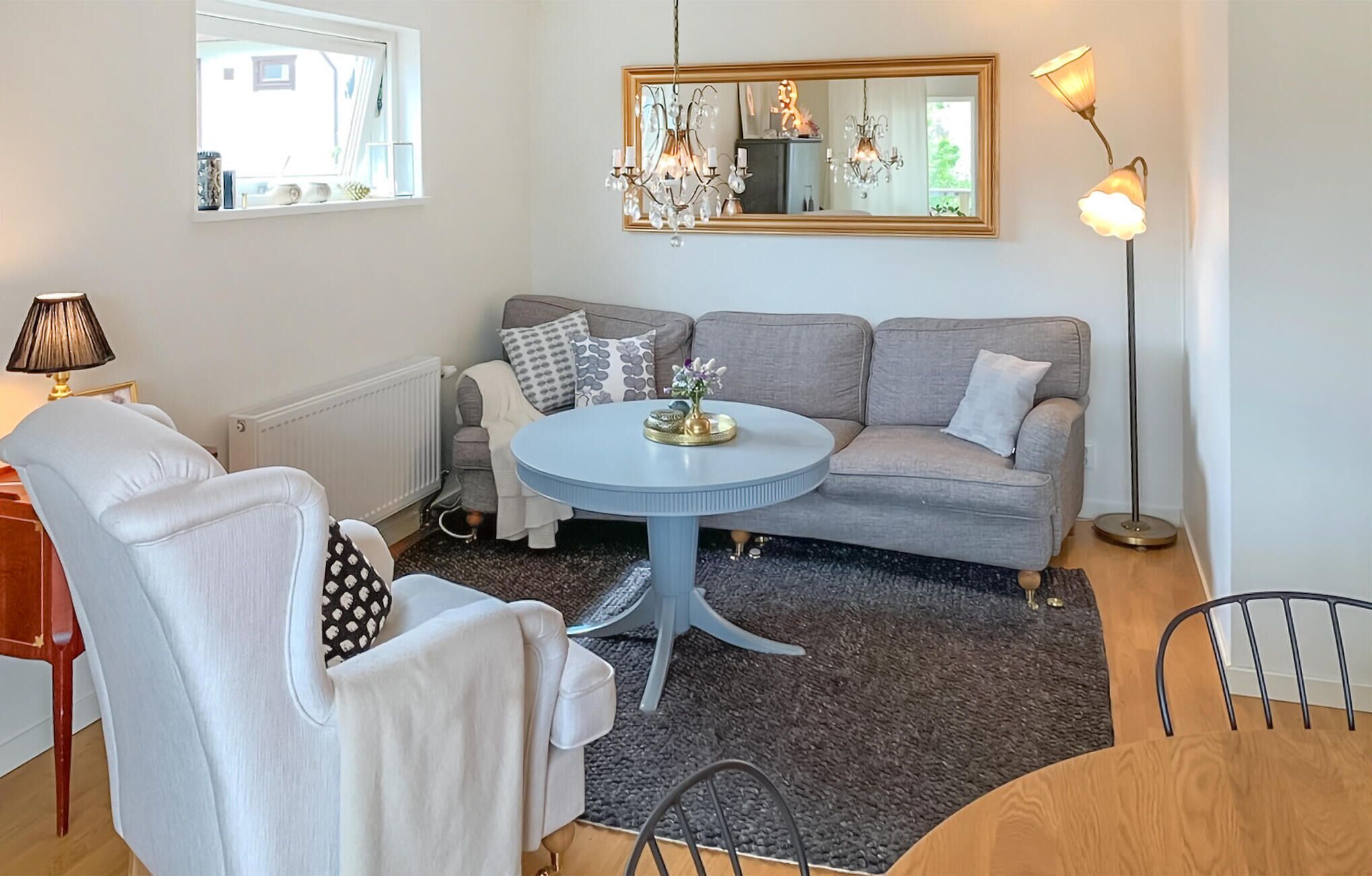 Un salon chaleureux avec un canapé gris, une table basse ronde et un fauteuil blanc. Une lampe sur pied et un miroir sont accrochés au mur.