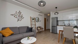 Un salon moderne avec un canapé gris, une petite table blanche et un fauteuil à motifs floraux. On aperçoit, au fond, une cuisine avec des meubles blancs et un réfrigérateur en acier inoxydable.