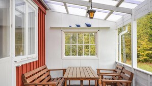 A covered outdoor seating area with wooden benches and a table, a window with a view of trees, and a lantern hanging above.
