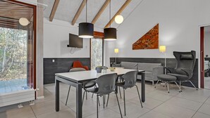 Et moderne spiseområde med et hvidt bord, sorte stole og en grå sofa. Pendellamper hænger over bordet, og der er et billede på væggen.