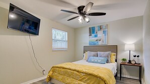 Une chambre à coucher comprenant un lit, un ventilateur de plafond, un téléviseur fixé au mur, une fenêtre et une table de chevet avec une lampe.