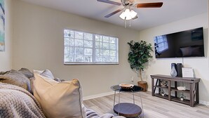 Un salon avec un ventilateur de plafond, une grande fenêtre, un téléviseur, une plante et une petite table.