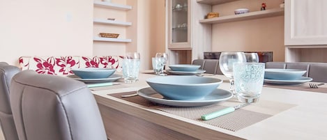 Uma sala de jantar com uma mesa comprida posta para quatro pessoas, com pratos azuis e copos de vidro transparente.