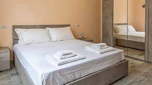 A neatly made bed with white linens and towels, a bedside table with a lamp, and a large mirror reflecting the room. - 2 bedroom amazing apartment in Guspini (Guspini)