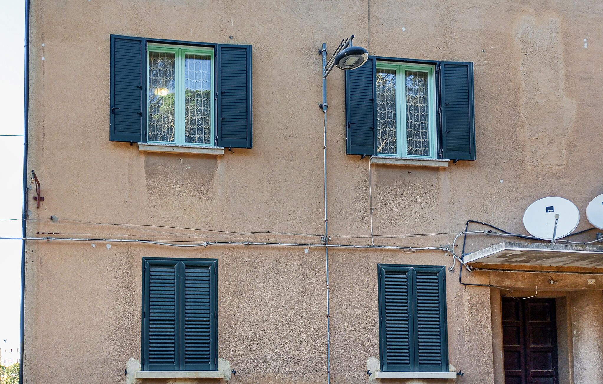 A building with three windows, each with green shutters. There are visible electrical wires and a satellite dish on the right side.